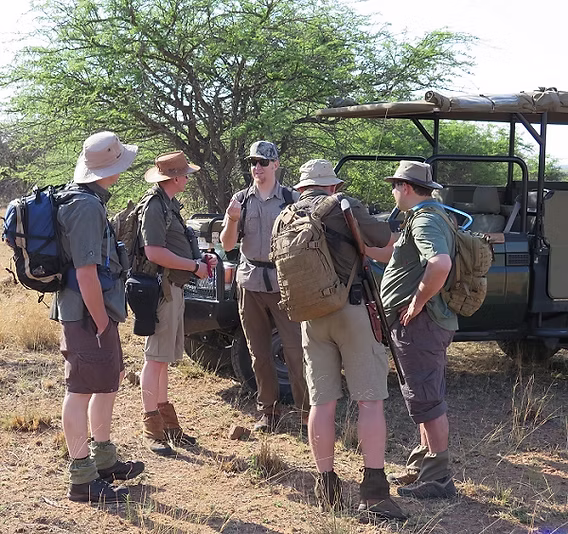 A safari traveler wearing neutral khaki and olive green clothing on an open game drive vehicle in the Masai Mara — correct safari clothing colors for a safari packing checklist